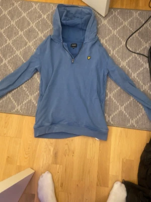 Blå hoodie från Lyle & Scott - Snygg blå hoodie från Lyle & Scott med halv dragkedja och klassisk gul logga på bröstet. Hoodien har en stor huva och ribbade muddar vid ärmslut och nederkant. Perfekt för en avslappnad och stilren look.
