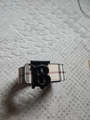 Rutmönstrat bälte med B-spänne - Snyggt bälte från Burberry med klassiskt rutmönster i beige, svart och rött samt ett stort svart B-spänne framtill. Bältet är gjort i skinn och har en exklusiv känsla. Perfekt accessoar för att lyfta din outfit. Den är dubbelsidig