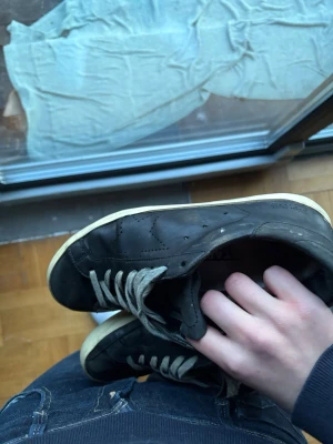 Svarta sneakers med vit sula - Svarta sneakers med vit sula och gråa skosnören. Klassisk låg modell med diskreta detaljer och perforeringar på sidan. Ovandelen är i läder eller syntetmaterial. Passar perfekt till streetwear och casual outfits. Skorna är i stort behov av hjälp där den är sönder på flera ställen.