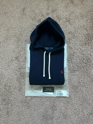 Mörkblå Ralph Lauren hoodie - En mörkblå Ralph Lauren Hoodie i storlek Medium. Den köptes ny på Boozt, och allt og medföljer vid köp.                                                    Den är i mycket fint skick, förutom en väldigt liten skada uppe vid luvan. Skadan är väldigt väldigt liten och placeringen gör att den knappt märks, då luvan täcker! Tveka inte på att höra av dig vid minsta fundering!⭐️🤙
