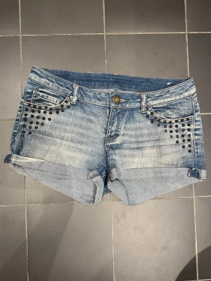 Blå jeansshorts med nitar från F3 Sister - Snygga blå lågmidjade jeansshorts från F3 Sister med slitna detaljer och svarta nitar vid fickorna framtill. Jag säljer shortsen eftersom de är för små för mig, men de är knappt använda och är i perfekt skick! Perfekta för sommaren och gör ens sommar outfits skitsnygga! Midjemått tvärs över: ca 37cm