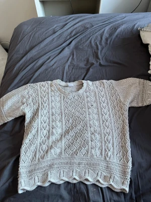 Grå stickad tröja med mönster - Snygg grå stickad tröja med unikt bladmönster och strukturstickade detaljer. Tröjan har rund halsringning, korta ärmar och bred ribbad kant nedtill. Perfekt för dig som gillar mysiga och stilrena plagg med lite extra detalj.