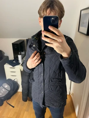 Mörkblå quiltad jacka Morris - Säljer en mörkblå quiltad jacka från Morris med klassisk krage och knappar framtill. Jackan har två stora fickor och ett emblem på ena ärmen. Perfekt för höst och vår, snygg och tidlös design som passar till jeans.