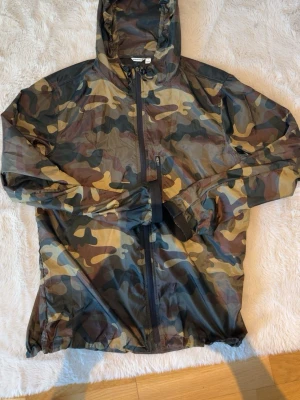 Kamo vindjacka med huva - Snygg vindjacka i camouflage-mönster med dragkedja framtill och huva. Jackan är lätt och perfekt för blåsiga dagar. Färgerna går i grönt, brunt och beige. Praktisk ficka med dragkedja på bröstet. Perfekt för våren PASSAR STORLEK S OCH M