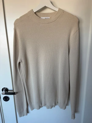 Beige stickad tröja med rund hals - En stilren beige stickad tröja med rund halsringning och ribbade muddar vid ärmslut och nederkant. Tröjan har en klassisk passform och är perfekt för lager-på-lager. Materialet ser ut att vara mjukt och bekvämt, vilket gör den enkel att matcha med olika outfits. Använd max 3 gånger på grund av att jag inte tyckte den passade mig. 