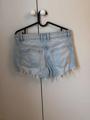 Ljusblå jeansshorts med fransar - Ljusblå jeansshorts med fransig kant. Shortsen har fickor, bälteshällor och knappstängning fram. Perfekta för varma dagar. Shortsen är använda men i gott skick.