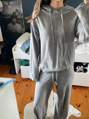 Grå loungemys-set med hoodie - Mysigt grått loungewear-set med hoodie och matchande byxor. Hoodien har dragsko och långa ärmar, byxorna har elastisk midja med snörning. Perfekt för chill dagar hemma. Materialet ser ut att vara mjukt och stretchigt.