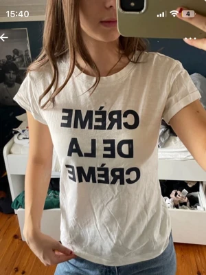 Vit t-shirt med svart text - Vit t-shirt med trycket 'CRÈME DE LA CRÈME' i svart. Klassisk rund hals och korta ärmar, perfekt för en avslappnad och cool look. Materialet känns mjukt och lätt, troligen bomull. Passformen är normal och t-shirten är enkel att matcha med jeans eller shorts.