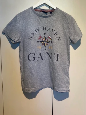 Grå t-shirt från GANT med tryck - Snygg grå t-shirt från GANT med stort tryck på bröstet där det står 'NEW HAVEN' och 'GANT' samt en kompassros och siffrorna 1949. Klassisk rund halsringning och korta ärmar. Perfekt för en avslappnad stil. Litet hål på ryggen men syns knappt
