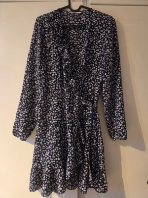 Blommig omlottklänning i marinblått - Söt marinblå kortklänning med vitt blommönster och omlottdesign. Klänningen har långa ärmar, volangkant och knytband i midjan. Perfekt för dig som gillar en feminin och avslappnad stil. Står storlek 34 men mer åt 36.