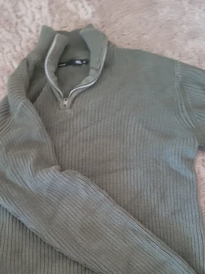 Olivgrön stickad halvzip från 157 - Olivgrön stickad tröja från 157 med halvzip och hög krage. Tröjan har ribbstickad struktur och långa ärmar, perfekt för lager på lager. Enkel och stilren design som passar till det mesta.