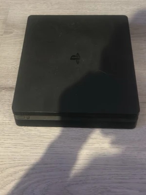 PlayStation 4 Slim svart konsol - Säljer en svart PlayStation 4 Slim från Sony. Konsolen har en stilren, tunn och fyrkantig design med mattsvart finish och PlayStation-logga på ovansidan. Utrustad med HDMI, USB och nätverksport på baksidan. Perfekt för gaming och streaming.