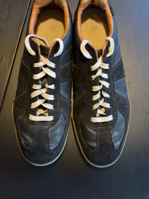 Svarta sneakers från H&M - Snygga svarta sneakers från H&M med vita skosnören och bruna detaljer på insidan. Skorna har en klassisk låg profil och är tillverkade i en mix av mocka och syntetmaterial. Perfekta för en avslappnad streetstyle-look.