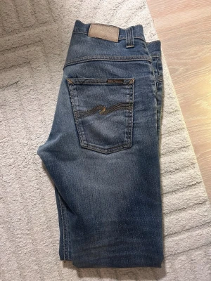 Blå jeans från Nudie Jeans - Säljer ett par klassiska blå jeans från Nudie Jeans med snyggt slitna detaljer och kontrastsömmar. Modellen har fem fickor och en smal passform. Materialet är mjukt denim i bomull och bakfickan har Nudie Jeans ikoniska brodyr. W33 men passar mer 30-31