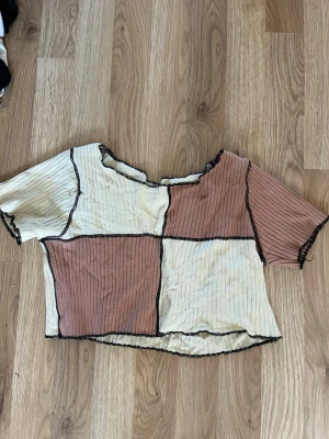 Beige och brun ribbad croppad topp - Trendig croppad topp i beige och brun med svarta kontrastsömmar. Toppen har korta ärmar, ribbad struktur och patchwork-design med synliga sömmar. Perfekt för en avslappnad och cool stil.