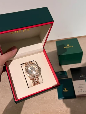 Role klocka i guld med diamanter - Exklusiv Role klocka i guld med silverfärgad urtavla och gnistrande diamanter runt boetten och på armbandet. Klassisk rund form och lyxig känsla, perfekt för dig som vill sticka ut med en riktigt snygg accessoar.
