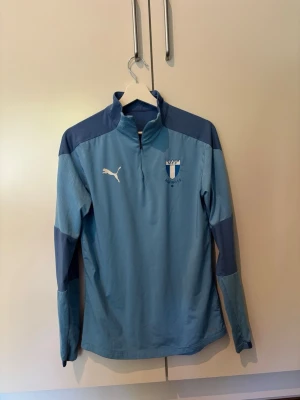 Ljusblå träningströja Malmö FF - Ljusblå långärmad träningströja från Puma med Malmö FF-logga på bröstet. Tröjan har dragkedja vid halsen, mörkare blå partier på axlar och ärmar samt vit Puma-logga. Perfekt för fotbollsträning eller sportiga aktiviteter. Storlek 154!