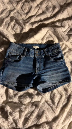 Blå denimshorts med medium midja - Säljer ett par klassiska blå denimshorts från “Denim Day?” med hög midja och fyra fickor. Shortsen har bälteshällor, gylf med dubbla knappar och bakfickor med lock. Perfekta för varma dagar och enkel att matcha.