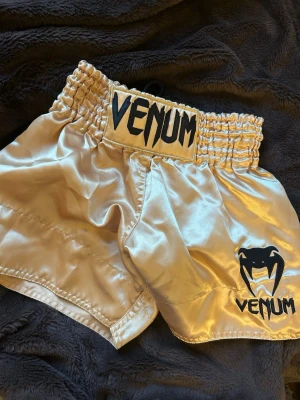 Guldiga thaishorts från Venum - Snygga guldiga thaiboxningsshorts från Venum med svart logga och resår i midjan. Tillverkade i glansigt, slitstarkt material som passar perfekt för träning eller match. Klassisk Venum-design med ormmotiv och bred midjeresår.