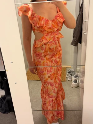 Lång blommig klänning med volanger - Storlek L men justerbar, köpt på vinted aldrig använd endast tvättad. Säljer en långklänning i orange, rosa och gul med blommigt mönster. Klänningen har volangdetaljer, öppen rygg med knyt och cut out i midjan. Ärmlös design med breda axelband och v-ringad hals. Perfekt för sommarens alla tillfällen, defekterna syns inte när man har på sig den.