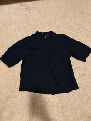 Mörkblå blus från H&M med knyt - En mörkblå blus från H&M med korta ärmar och dekorativt knytband vid halsen. Blusen har en avslappnad passform och smockdetaljer över bysten. Tillverkad i ett lätt och luftigt tyg, perfekt för en trendig och bekväm stil.
