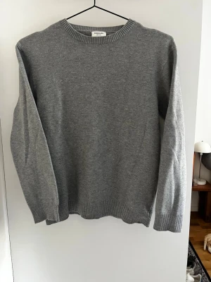 Grå stickad tröja från Kappahl - Klassisk grå stickad tröja från Kappahl med rund halsringning och ribbade muddar vid ärmslut och nederkant. Enkel och stilren design som passar till det mesta. Tillverkad i mjukt material för skön känsla.