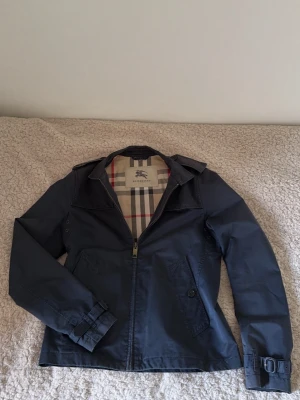 Vintage Burberry - Snygg mörkblå vindjacka från Burberry med klassiskt rutigt foder. Jackan har dragkedja framtill, axelklaffar och spännen vid ärmsluten. Perfekt för dig som vill ha en stilren och tidlös jacka med exklusiv känsla.