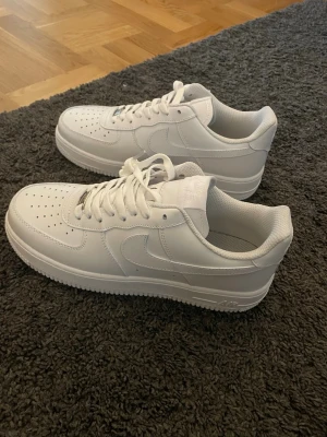 Nike Air Force 1 vita sneakers - Klassiska Nike Air Force 1 sneakers i helvitt läder med perforerad tåbox, vit sula och vita skosnören. Ikonisk swoosh på sidan och metallbricka med 'AF1' på snörningen. Perfekt för dig som gillar stilrena och tidlösa sneakers.