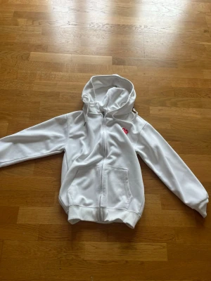 Vit hoodie från Comme des Garçons Play - Vit hoodie med dragkedja från Comme des Garçons Play. Klassisk modell med huva och fickor framtill. På bröstet finns det ikoniska röda hjärtat med ögon. Mjuk bomullskänsla och stilren design som passar perfekt till streetwear.