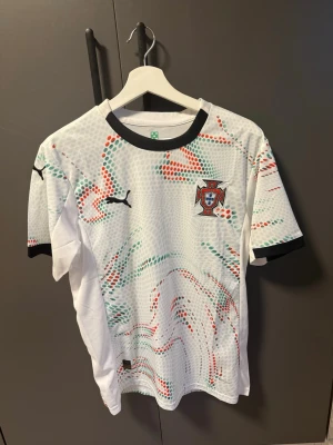Portugal fotbollströja Puma vit - Vit Portugal fotbollströja från Puma med svart krage och ärmslut. Tröjan har färgglada prickmönster i grönt och rött samt Portugals landslagsemblem på bröstet. Perfekt för fotbollsträning eller att bära när du hejar på laget.