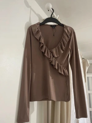 Brun omlottopp med volang - Snygg brun omlottopp med volangdetaljer längs ringningen. Långärmad modell i mjukt material, perfekt för att lyfta en enkel outfit. Stilren och trendig design som passar till många tillfällen.