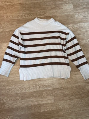 Randig stickad tröja i vitt och brunt - Stickad tröja med bruna och vita ränder. Tröjan har ribbstickade muddar, slits i sidorna och en avslappnad passform. I storlek M
