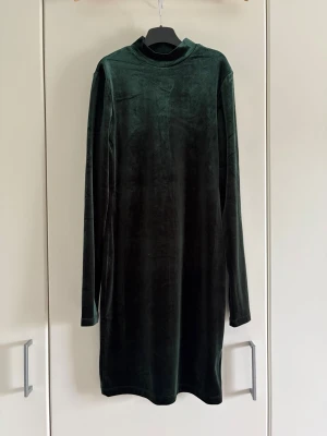 Mörkgrön sammetsklänning med polokrage - Mörkgrön sammetsklänning med lång ärm och polokrage. Klänningen har en rak passform och slutar ovanför knäna. Perfekt för dig som gillar en stilren och elegant look med en mjuk känsla.