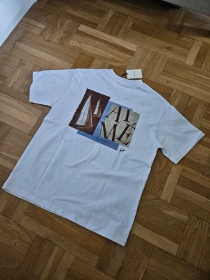 Vit Aimé Leon Dore t-shirt med tryck - Säljer en vit t-shirt från Aimé Leon Dore med ett stort grafiskt tryck på ryggen som visar segelbåtar och texten 'AIME'. T-shirten har klassisk passform och korta ärmar. Perfekt för dig som gillar stilrena plagg med unik design.