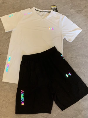 Under Armour sportset i vitt och svart - Snyggt sportset från Under Armour med en vit t-shirt och svarta shorts. Setet har holografiska loggor och textdetaljer i regnbågsfärger. Tillverkat i ett lätt och snabbtorkande material som passar perfekt till träning eller gym.