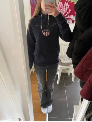 Mörkblå hoodie från GANT med logga - Mörkblå hoodie från GANT med stor broderad logga och amerikanskinspirerat sköldmärke på bröstet. Klassisk huva med vita dragsnören och känguruficka framtill. Perfekt för en avslappnad och stilren look.