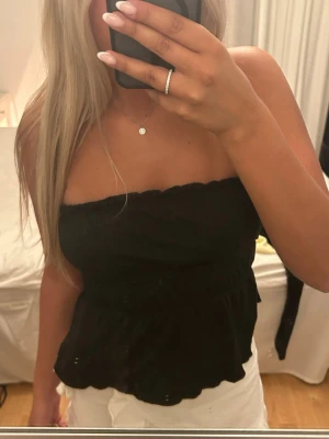 Svart bandeau tubtopp - Säljer en svart bandeau tubtopp med volangkant nertill. Toppen är axelbandslös och har en smockad midja som ger en snygg passform. Perfekt till sommaren och enkel att matcha med olika stilar.