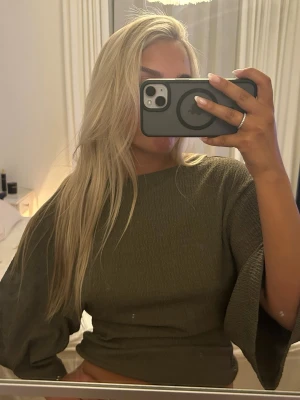 Olivgrön croppad topp med vida ärmar - Trendig olivgrön croppad topp med vida, trekvartsärmar och rund halsringning. Tillverkad i ett mjukt, ribbat material som ger en avslappnad men snygg look. Perfekt att styla med höga jeans eller kjol.