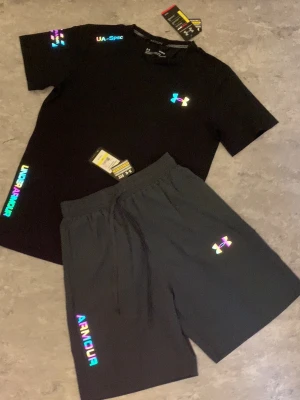 Svart träningsset från Under Armour - Snyggt svart träningsset från Under Armour med t-shirt och shorts. Setet har färgglada, reflekterande loggor och textdetaljer. T-shirten har rund hals och korta ärmar, shortsen har resår i midjan och dragsko. Perfekt för gymmet eller löprundan.