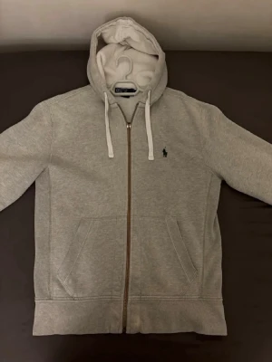 Grå hoodie från Polo Ralph Lauren - Snygg grå hoodie från Polo Ralph Lauren med dragkedja framtill, två fickor och klassisk huva med vita snören. Liten grön logga broderad på bröstet. Tillverkad i mjukt sweatshirtmaterial som är perfekt för chill dagar. Den är minimalt skadad i ärmarna och dragkedjan är lite skadad som man kan se på bilderna