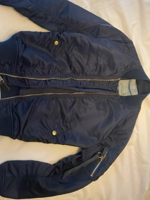 Marinblå bomberjacka Alpha Industries - Snygg marinblå bomberjacka från Alpha Industries med klassisk gulddragkedja, ribbade muddar och fickor med tryckknappar. Jackan har en ärmficka med dragkedja och ikonisk Alpha-tag. Tillverkad i glansigt nylonmaterial som ger en cool look.