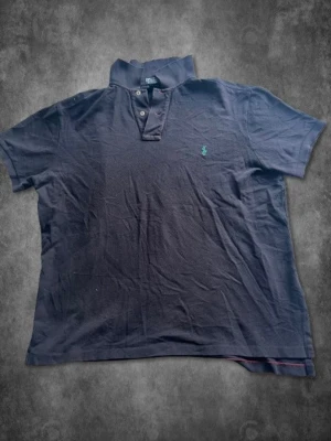 Mörkblå piké från Polo Ralph Lauren - Klassisk mörkblå pikétröja från Polo Ralph Lauren i 100% bomull. Tröjan har korta ärmar, knappar vid halsen och den ikoniska gröna logotypen broderad på bröstet. Perfekt för en avslappnad men ändå stilren look. Mått: Axelbredd: 47cm Pit-to-Pit: 57cm Midja: 53cm Längd: 63cm (räknar inte med krage)