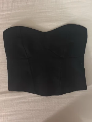 Svart korsett-topp  - Snygg svart korsett-topp med hjärtformad urringning och dragkedja bak. Toppen är axelbandslös och har en figurnära passform som framhäver midjan. Perfekt att styla med höga jeans eller kjol för en trendig look.
