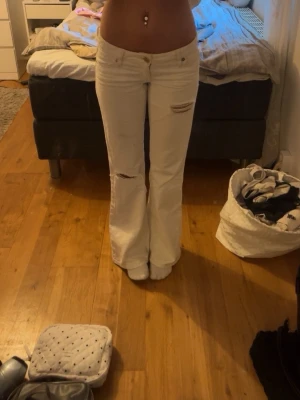 Vita bootcut jeans med slitningar - Säljer ett par vita bootcut jeans med slitningar på låren och knäna. Jeansen har hög midja och klassisk femficksdesign. Perfekta för dig som vill ha en trendig och avslappnad look.