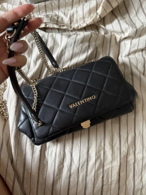Svart quiltad handväska från Valentino - Snygg svart handväska från Valentino med quiltat mönster och guldfärgade detaljer. Väskan har kedjerem och praktiskt innerfack med dragkedja. Perfekt accessoar för att lyfta din outfit.