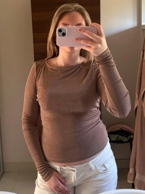 Brun långärmad topp i genomskynligt material - Mjuk och stilren brun långärmad topp i genomskynligt material. Enkel design med rund halsringning och rak passform. Perfekt att matcha med jeans eller kjol för en clean look.