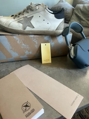 Golden Goose vita sneakers med stjärna - Golden Goose sneakers i vitt läder med grå stjärndetalj på sidan och svart häl. Skorna har en klassisk låg siluett, slitna kanter för en cool look och grå skosnören. Perfekta för dig som gillar streetstyle och exklusiva märken. OBS OSÄKER PÅ ÄKTHET!!
