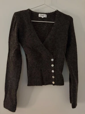 Mörkbrun kofta från Gina Tricot - Säljer en mörkbrun stickad kofta från Gina Tricot i alpackablandning. Koftan har djup V-ringning, långa ärmar och fem silvriga knappar framtill. Sitter tajt i midjan.