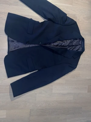 Svart kostym med kavaj och byxor - Stilren svart kostym med klassisk kavaj och matchande byxor. Kavajen har två knappar och fickor framtill, samt slät insida. Perfekt för dig som vill ha en tidlös och snygg look. Passar till många olika tillfällen. Kavaj: 44 short, slim fit. Byxor: 28-32