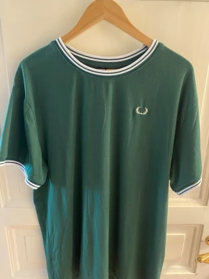 Grön Fred Perry t-shirt med vit logga - Snygg grön t-shirt från Fred Perry med vit broderad logga på bröstet. T-shirten har vita och blå ränder på krage och ärmslut, klassisk passform och är tillverkad i mjuk bomull. Perfekt för en avslappnad och stilren look.